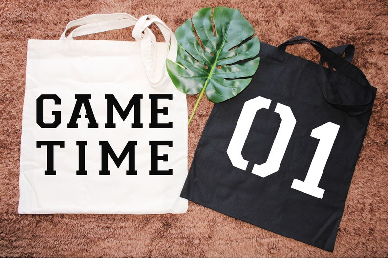 K&ouml;nnte beinhalten: Zwei wei&szlig;e Canvas-Tragetaschen mit schwarzem Text. Eine Tasche sagt "GAME TIME" und die andere sagt "01".