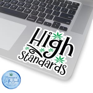 May include: A white sticker with the text "High Standards" in a black, stylized font, accented with green cannabis leaves. The sticker is on a laptop. A circular logo with the text "Flowers On The Beach" is in the lower left corner.