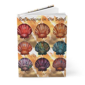 以下が含まれることがあります： 「Reflections in the Sand」というタイトルのハードカバーのジャーナル。表紙には、赤、オレンジ、緑、青、紫の色合いのカラフルな貝殻の模様が、金とベージュの背景に描かれています。