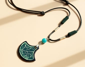 May include: A black cord necklace with a green and white ceramic pendant shaped like a fan. The pendant has a turquoise bead and silver accents.