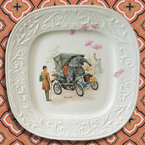 Könnte beinhalten: Ein quadratischer Teller aus weißem Keramik mit einem verzierten Rand. In der Mitte des Tellers befindet sich eine Vintage-Illustration eines Mannes, der neben einem Auto mit einem Elefanten am Steuer steht. Das Auto ist mit "Fiat 1901" beschriftet.