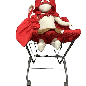 May include: A red hooded cape with a mask, worn by a mannequin sitting in a shopping cart. The cape has a pouch attached to the front.