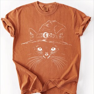 Rust orange garment-dyed tee laid flat, printed with a hand-drawn black cat wearing a moon buckle witch hat.