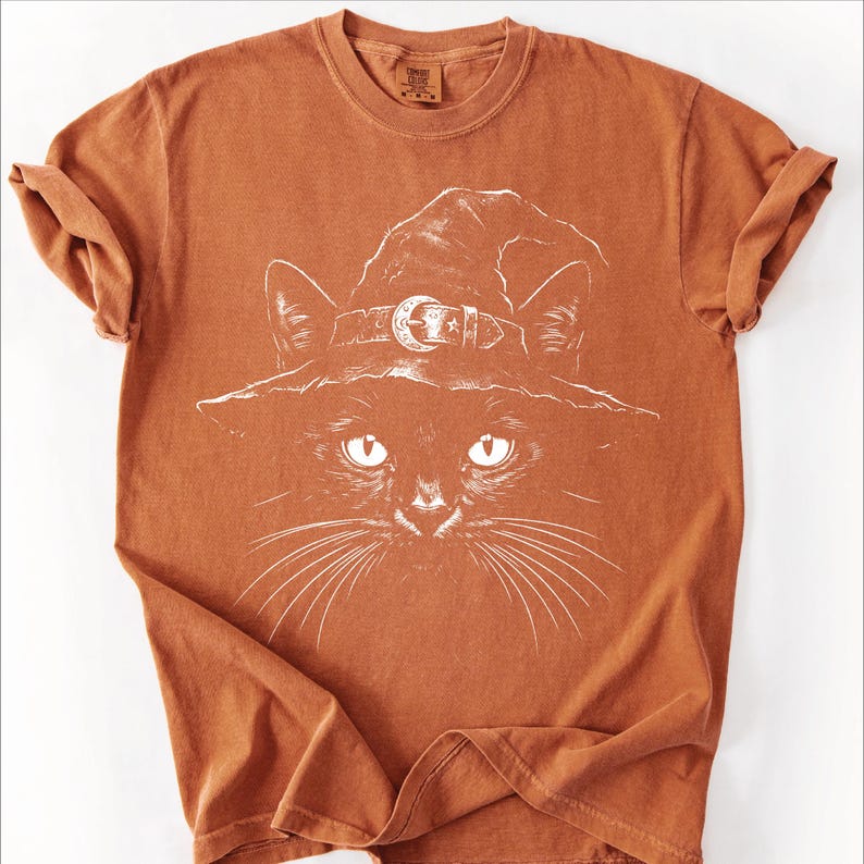 Rust orange garment-dyed tee laid flat, printed with a hand-drawn black cat wearing a moon buckle witch hat.