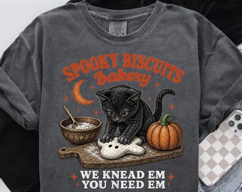 May include: A charcoal gray t-shirt featuring a graphic of a black kitten kneading dough on a wooden board. The design includes an orange crescent moon, a pumpkin, a mixing bowl, and the text: SPOOKY BISCUITS bakery WE KNEAD EM YOU NEED EM.