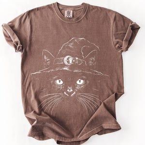 Warm brown garment-dyed t-shirt laid flat, showing the fine white line drawing of a black cat in a pointed witch hat.