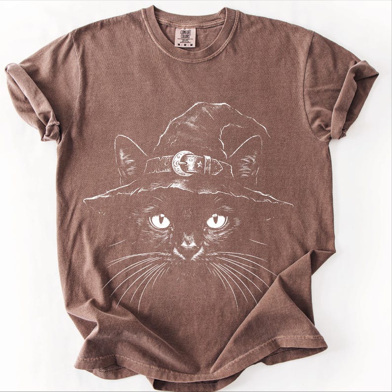 Warm brown garment-dyed t-shirt laid flat, showing the fine white line drawing of a black cat in a pointed witch hat.