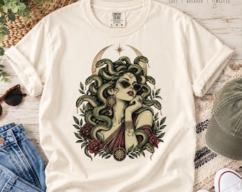 Könnte beinhalten: Ein cremefarbenes T-Shirt mit einem Grafikdruck einer Figur mit Schlangen als Haaren, die eine Sonnenbrille trägt, umgeben von einem Halbmond, Blättern und Blumen. Das T-Shirt hat ein Comfort Colors-Etikett am Hals mit dem Text Comfort Colors Soft Relaxed Timeless.