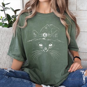 Model seated in an oversized moss green t-shirt featuring a detailed white black cat witch hat illustration.