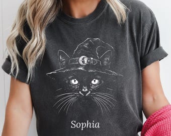 May include: A charcoal gray short-sleeve t-shirt featuring a white line art graphic of a cat wearing a witch hat with a crescent moon buckle. The name Sophia is printed in white text below the cat illustration on the front of the garment.