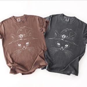 Two garment-dyed tees side by side in brown and dark gray, both printed with the same white black cat witch hat design.