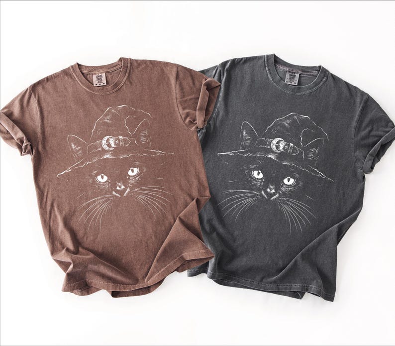 Two garment-dyed tees side by side in brown and dark gray, both printed with the same white black cat witch hat design.