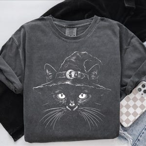 Folded dark gray garment-dyed t-shirt with a white line-art black cat in a witch hat, styled flat with denim and a phone.