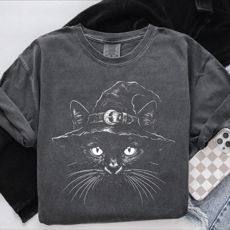 Folded dark gray garment-dyed t-shirt with a white line-art black cat in a witch hat, styled flat with denim and a phone.