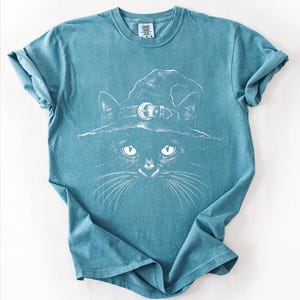Teal blue Comfort Colors 1717 t-shirt flat lay with a white witchy black cat illustration printed on the front.