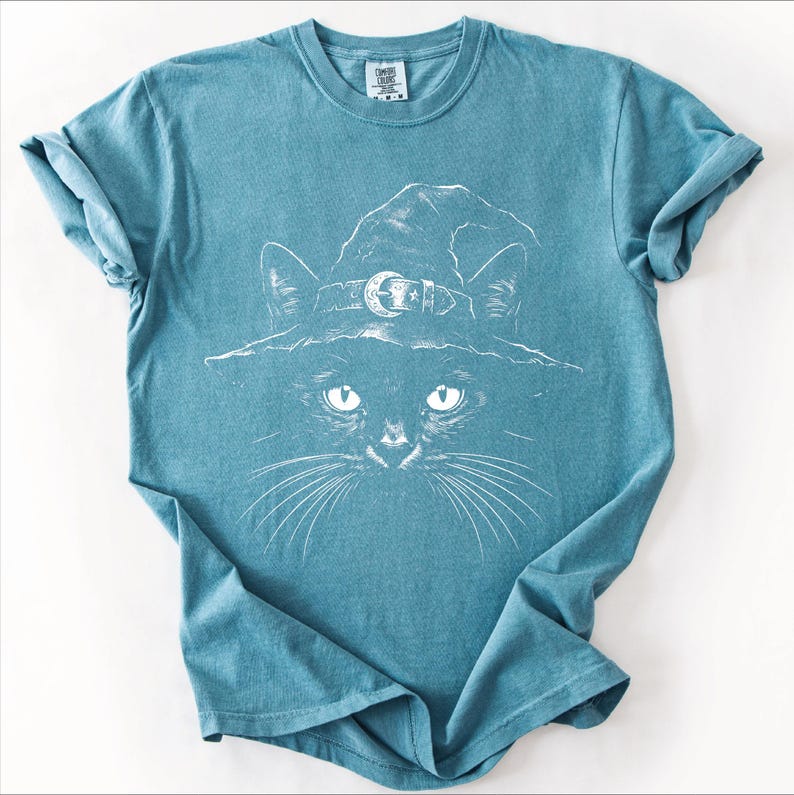 Teal blue Comfort Colors 1717 t-shirt flat lay with a white witchy black cat illustration printed on the front.