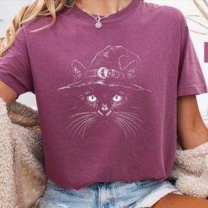 Woman wearing a deep berry Comfort Colors tee printed with a hand-drawn black cat in a crescent moon witch hat.