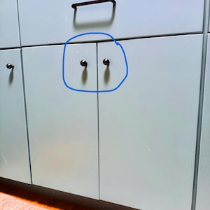 Pode incluir: Close-up de uma seção de um armário azul claro com ferragens pretas. As portas do armário têm pequenos botões pretos redondos. Uma alça preta retangular está acima das portas do armário.