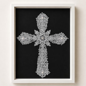Könnte beinhalten: Ein gerahmtes Wandkunstwerk mit einem Kreuz aus filigranem, silberfarbenem Papier-Quilling. Das Kreuz besteht aus zahlreichen engen Spiralen und blattförmigen Wicklungen, akzentuiert mit kleinen weißen Perlen auf schwarzem Hintergrund.
