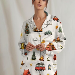 以下が含まれることがあります： キャンプをテーマにした白いパジャマセット。ボタンダウンシャツとマッチするパンツのセットで、テント、キャンプファイヤー、木、ヴィンテージカーのカラフルなイラストが描かれています。シャツとパンツの縁は黒いパイピングでトリミングされています。