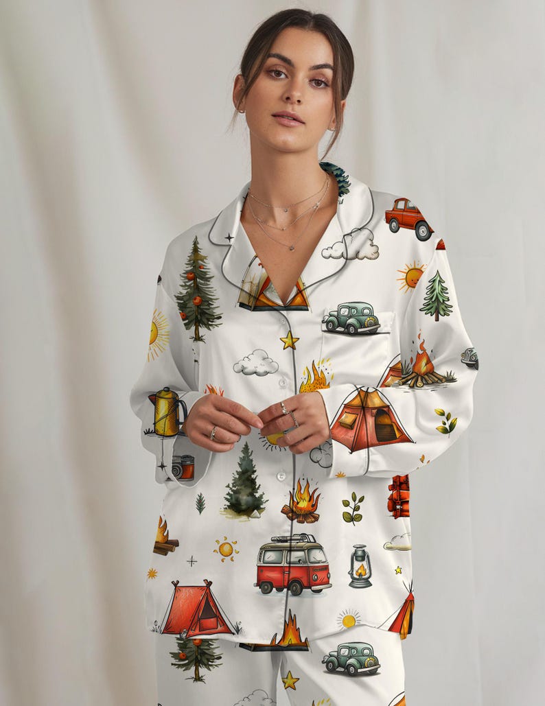 以下が含まれることがあります： キャンプをテーマにした白いパジャマセット。ボタンダウンシャツとマッチするパンツのセットで、テント、キャンプファイヤー、木、ヴィンテージカーのカラフルなイラストが描かれています。シャツとパンツの縁は黒いパイピングでトリミングされています。