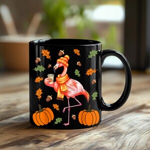 May include: Black ceramic mug featuring a flamingo wearing a knit hat and scarf, holding a coffee cup. The mug is decorated with pumpkins, autumn leaves, and acorns. A perfect fall-themed gift.