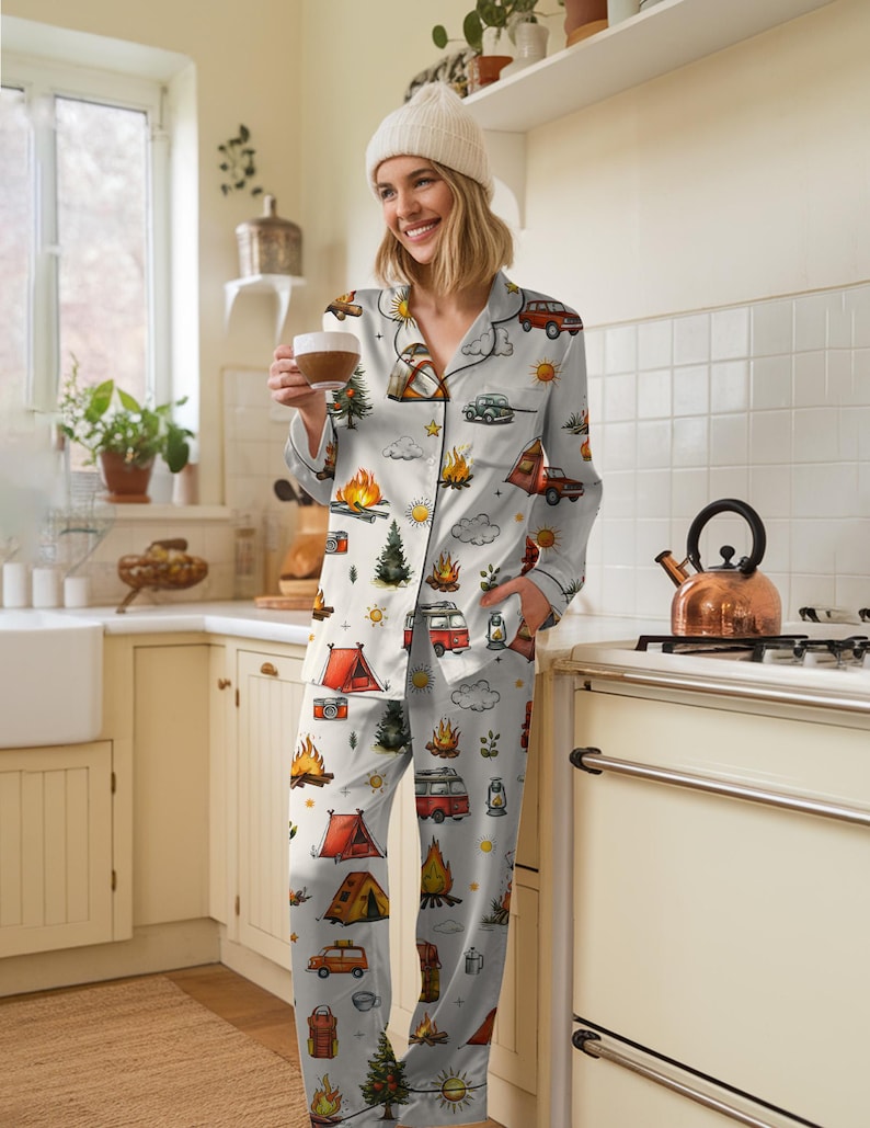 以下が含まれることがあります： キャンプをテーマにしたライトグレーのパジャマセット。ボタンダウンシャツとパンツのセットで、テント、キャンプファイヤー、木、ヴィンテージキャンピングカーのイラストが描かれています。パジャマは柔らかい素材で作られています。