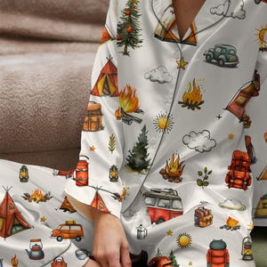以下が含まれることがあります： キャンプをテーマにした白いパジャマセット。テント、キャンプファイヤー、木、車、バックパック、ランタンのイラストが特徴です。デザインにはオレンジ、赤、緑、黄色のアクセントが含まれています。パジャマは、襟付きのボタンダウンのトップスとマッチングパンツです。