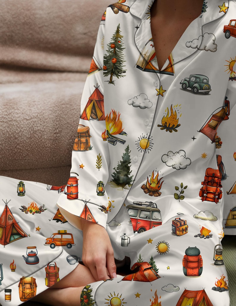 以下が含まれることがあります： キャンプをテーマにした白いパジャマセット。テント、キャンプファイヤー、木、車、バックパック、ランタンのイラストが特徴です。デザインにはオレンジ、赤、緑、黄色のアクセントが含まれています。パジャマは、襟付きのボタンダウンのトップスとマッチングパンツです。