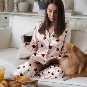 K&ouml;nnte beinhalten: Eine Person in einem rosa Pyjama mit Weinglasmuster, die eine schwarze Tasse und ein Buch h&auml;lt, sitzt mit einem kleinen Hund auf einem wei&szlig;en Sofa. Ein Tablett mit Croissants und Saft steht auf einem Couchtisch.