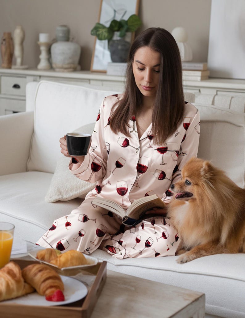 K&ouml;nnte beinhalten: Eine Person in einem rosa Pyjama mit Weinglasmuster, die eine schwarze Tasse und ein Buch h&auml;lt, sitzt mit einem kleinen Hund auf einem wei&szlig;en Sofa. Ein Tablett mit Croissants und Saft steht auf einem Couchtisch.