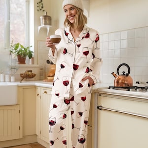 K&ouml;nnte beinhalten: Cremefarbenes Pyjama-Set mit einem sich wiederholenden Muster aus roten Weingl&auml;sern. Das lang&auml;rmlige Oberteil und die Hose haben schwarze Paspeln. Die Pyjamas sind aus seidigem Material. Die Person im Bild tr&auml;gt eine wei&szlig;e M&uuml;tze.