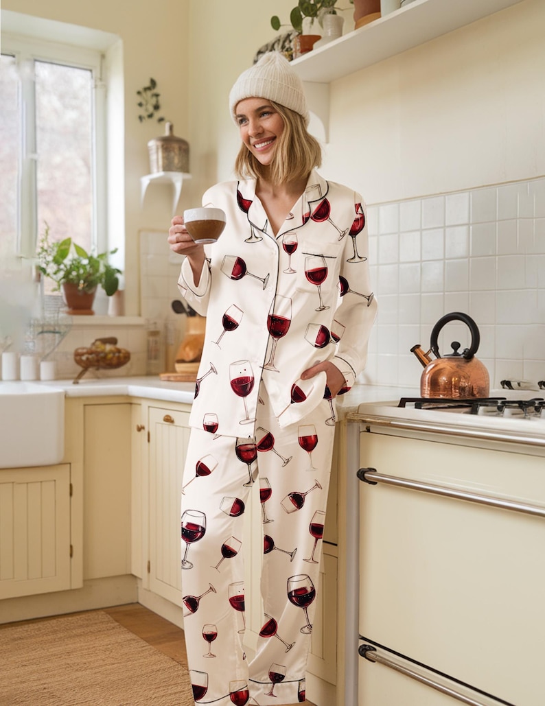 K&ouml;nnte beinhalten: Cremefarbenes Pyjama-Set mit einem sich wiederholenden Muster aus roten Weingl&auml;sern. Das lang&auml;rmlige Oberteil und die Hose haben schwarze Paspeln. Die Pyjamas sind aus seidigem Material. Die Person im Bild tr&auml;gt eine wei&szlig;e M&uuml;tze.