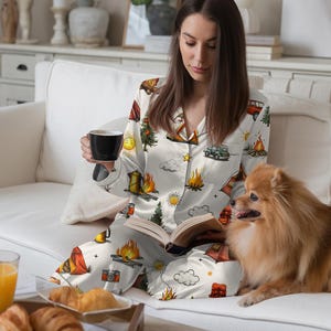 以下が含まれることがあります： テント、キャンプファイヤー、木、ヴィンテージカーが描かれた、キャンプをテーマにしたプリントの白いパジャマセット。 セットには、ボタンダウンシャツとパンツが含まれています。 白いソファに座って本を読み、黒いマグカップを持っている人がいます。 小さな犬がその人の隣に座っています。