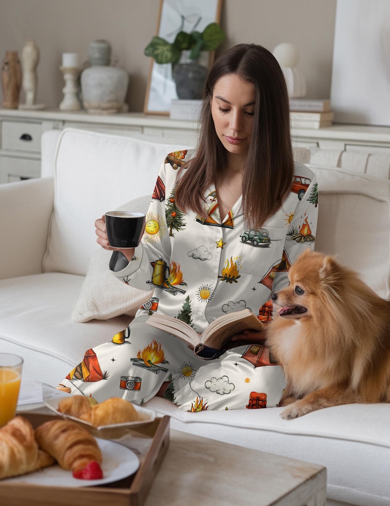 以下が含まれることがあります： テント、キャンプファイヤー、木、ヴィンテージカーが描かれた、キャンプをテーマにしたプリントの白いパジャマセット。 セットには、ボタンダウンシャツとパンツが含まれています。 白いソファに座って本を読み、黒いマグカップを持っている人がいます。 小さな犬がその人の隣に座っています。