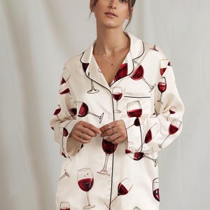 K&ouml;nnte beinhalten: Cremefarbenes Pyjama-Set mit einem sich wiederholenden Muster aus roten Weingl&auml;sern. Das Set beinhaltet ein Hemd mit Kn&ouml;pfen und schwarzem Besatz sowie eine passende Hose. Der Pyjama ist aus einem weichen, seidigen Material gefertigt.