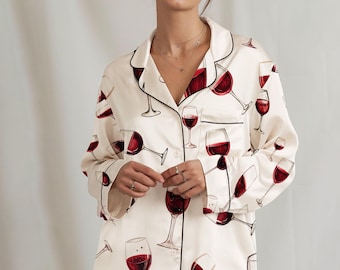 May include: Cream-colored pajama set featuring a repeating pattern of red wine glasses. The set includes a button-down shirt with black trim and matching pants. The pajamas are made of a soft, silky material.