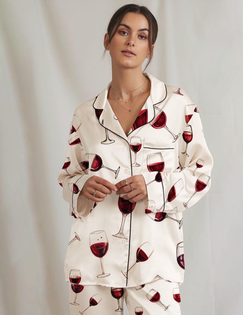 K&ouml;nnte beinhalten: Cremefarbenes Pyjama-Set mit einem sich wiederholenden Muster aus roten Weingl&auml;sern. Das Set beinhaltet ein Hemd mit Kn&ouml;pfen und schwarzem Besatz sowie eine passende Hose. Der Pyjama ist aus einem weichen, seidigen Material gefertigt.