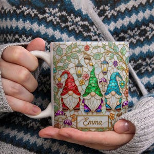 May include: White ceramic mug featuring a stained-glass design with three gnomes wearing red, green, and blue hats. The mug includes the name "Emma" and decorative elements like lanterns, flowers, and mushrooms. The mug is held in a person's hands.
