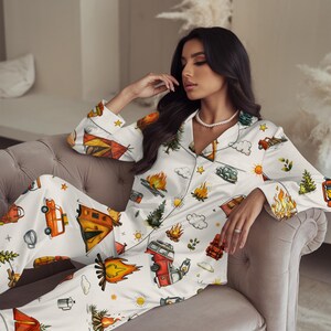 以下が含まれることがあります： キャンプをテーマにした白いパジャマセット。ボタンダウンのトップスとパンツがあり、オレンジ、赤、緑色のキャンプファイヤー、テント、木、ヴィンテージキャンピングカーのイラストが描かれています。パジャマはシルクのような素材で作られています。