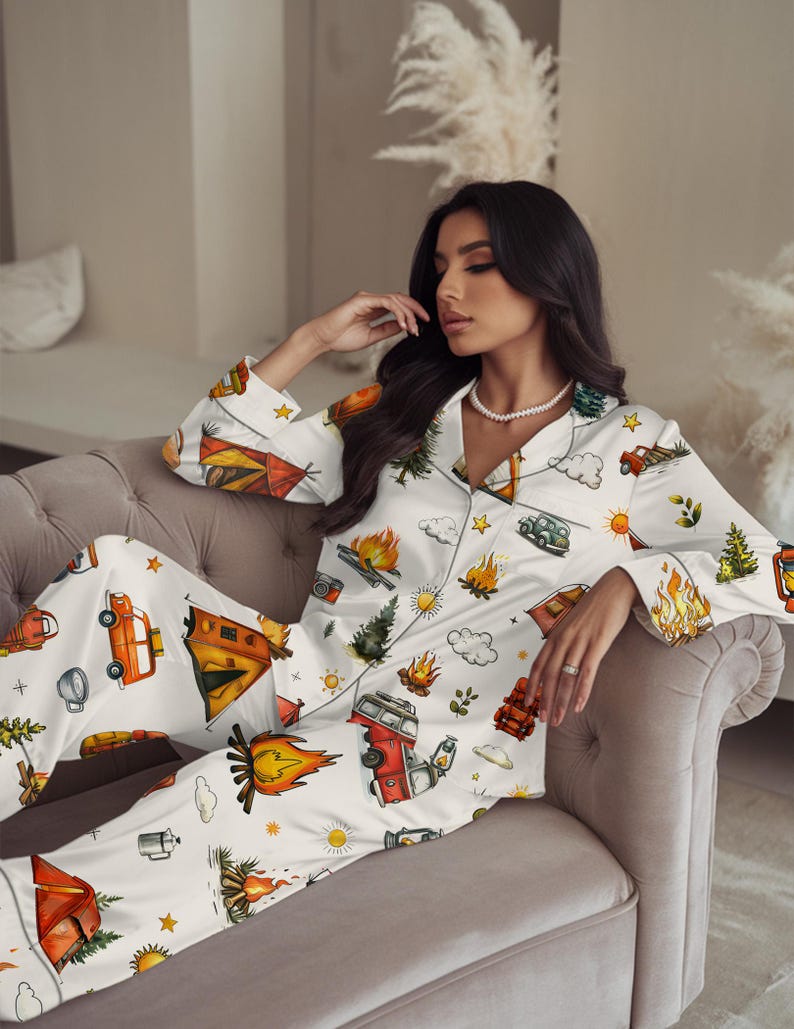 以下が含まれることがあります： キャンプをテーマにした白いパジャマセット。ボタンダウンのトップスとパンツがあり、オレンジ、赤、緑色のキャンプファイヤー、テント、木、ヴィンテージキャンピングカーのイラストが描かれています。パジャマはシルクのような素材で作られています。