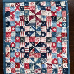 May include: A square, handmade patchwork quilt with a blue border. The quilt is composed of small, colorful fabric squares in red, white, and blue, with various patterns including polka dots, stripes, and floral designs. The quilt has a nautical theme.