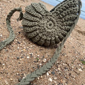 May include: An olive green crocheted bag shaped like a nautilus shell, featuring a spiraled texture and a thick, braided shoulder strap. The bag is constructed from thick yarn and includes gold-toned metal rings connecting the strap to the body.