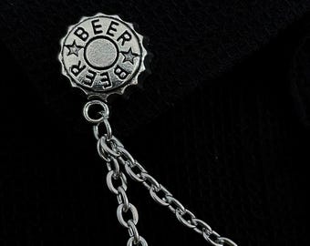 Può includere: Spilla da bavero color argento a forma di tappo di birra, con la scritta "BEER" e stelle. La spilla è attaccata a una doppia catena, creando un accessorio elegante. Lo sfondo è nero.