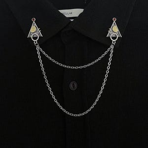 Peut inclure: Une chaîne de col argentée avec deux broches triangulaires. Chaque broche présente un motif doré et une gemme rouge. La chaîne est suspendue entre les broches, fixées à une chemise noire.