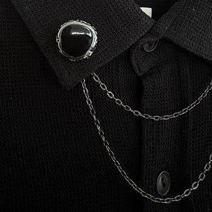 Puede incluir: Primer plano de una camisa negra con tejido texturizado. La camisa presenta una cadena plateada decorativa y dos botones negros. El cuello tiene un botón negro grande y redondo con borde plateado.