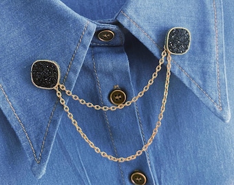 May include: A gold chain collar pin with two black square-shaped stones. The pin is attached to a denim shirt.