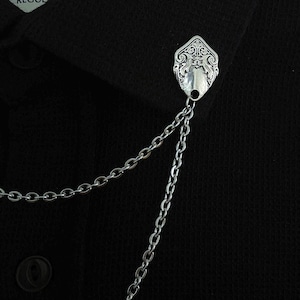 May include: A silver-toned metal collar pin featuring an ornate, shield-shaped emblem with intricate embossed scrollwork patterns. Two delicate silver-toned link chains hang from the base of the emblem, attached to a black textured fabric.