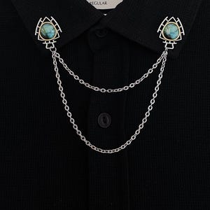 以下が含まれることがあります： 2つの装飾ピンが付いたシルバーカラーの襟チェーン。各ピンは、金色のフレームと青緑色の石が特徴です。チェーンは黒い襟付きシャツの上にドレープされています。