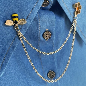 Peut inclure: Broche dorée en forme d'abeille et de nid d'abeilles avec une chaîne, attachée à une chemise en jean bleue. L'abeille a des détails en émail noir et jaune. Le nid d'abeilles a des détails en émail orange et marron.